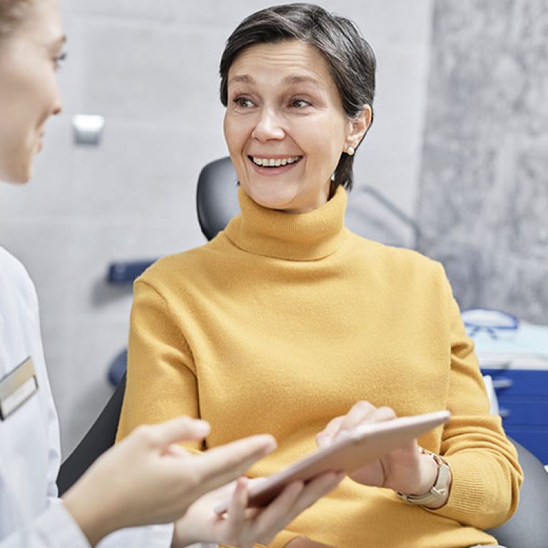 uniquedent_web_services_0007_smiling-mature-woman-in-dental-clinic-talking-to-f-2026-01-08-06-58-15-utc